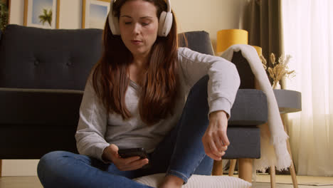 Mujer-Con-Auriculares-Inalámbricos-Sentada-En-El-Suelo-En-Casa-Transmitiendo-Música-Desde-Un-Teléfono-Móvil-3